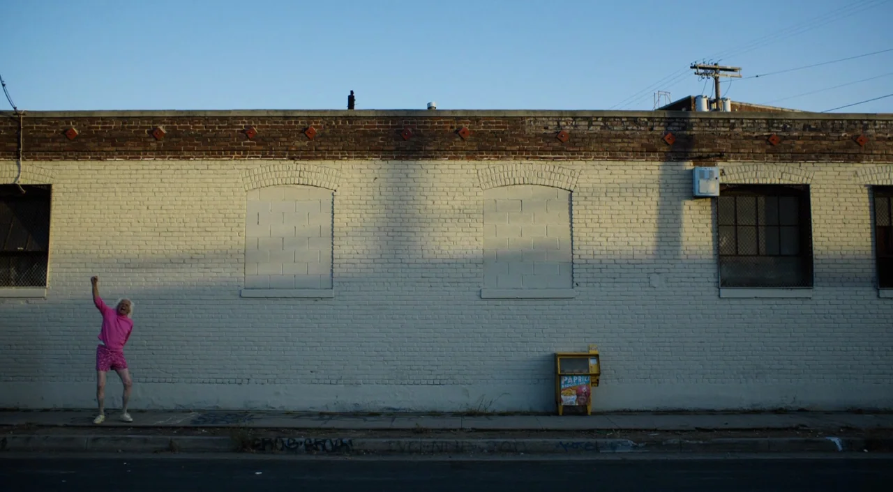 The Greasy Strangler (2016) movie still: wide — A long, single-story industrial building features white-painted brick and blocked-up window…