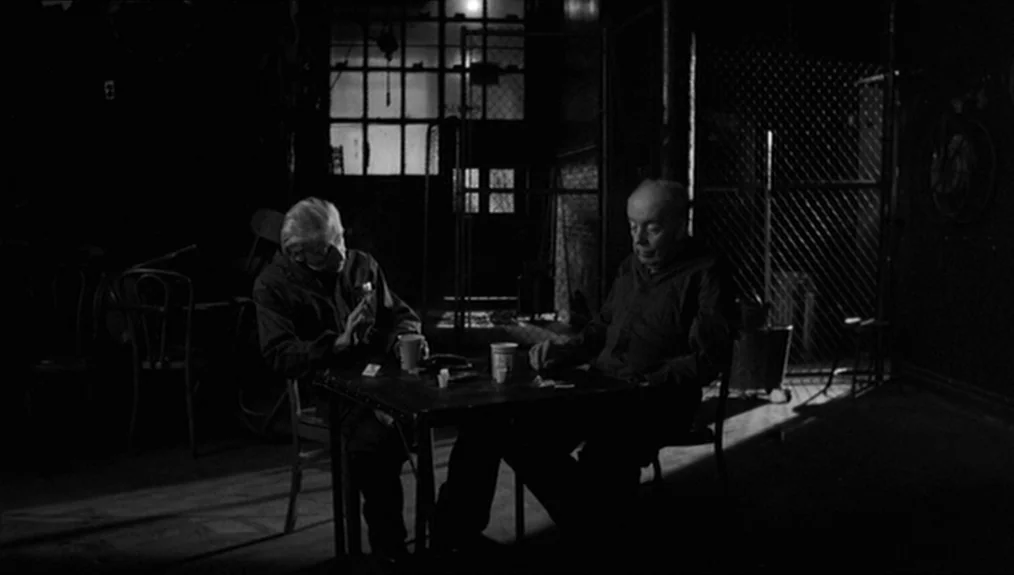Coffee and Cigarettes (2003) movie still: medium — A dark, spartan room features a chain-link fence partition and large industrial window…