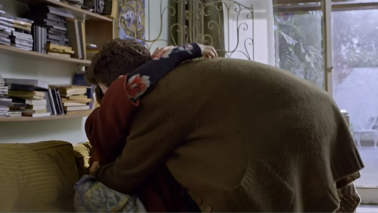 Heaven Sent (1994) movie still: medium — A residential living room contains a bookshelf, a couch, and a glass door window. Two individuals embrace…