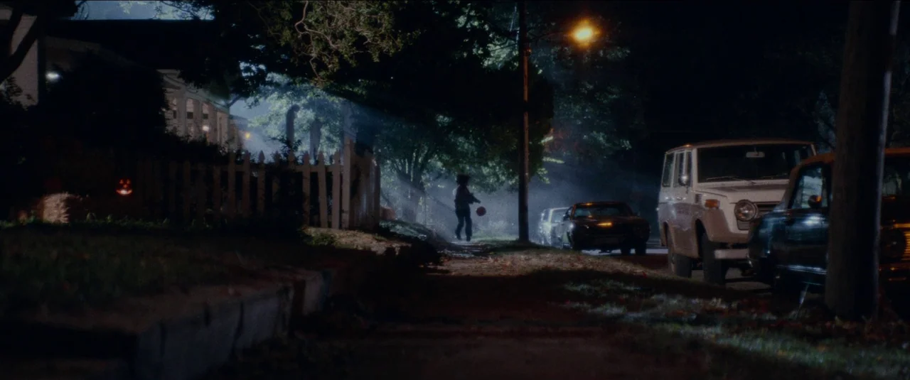 Halloween Kills (2021) movie still: wide — A dark residential street is blanketed in thick fog under the night sky. A lone child walks down the…