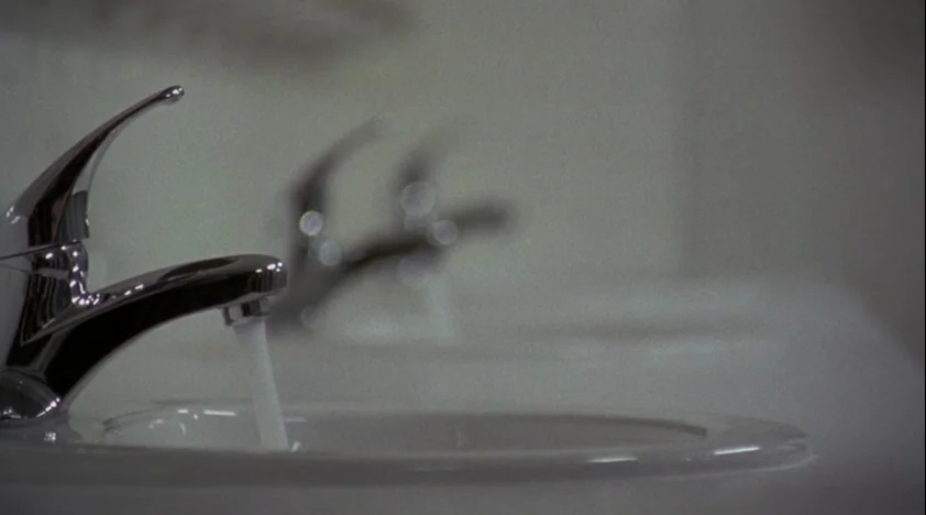The House Of The Devil (2009) movie still: closeup — A close-up view of a polished chrome faucet over a white porcelain basin. A stream of water…