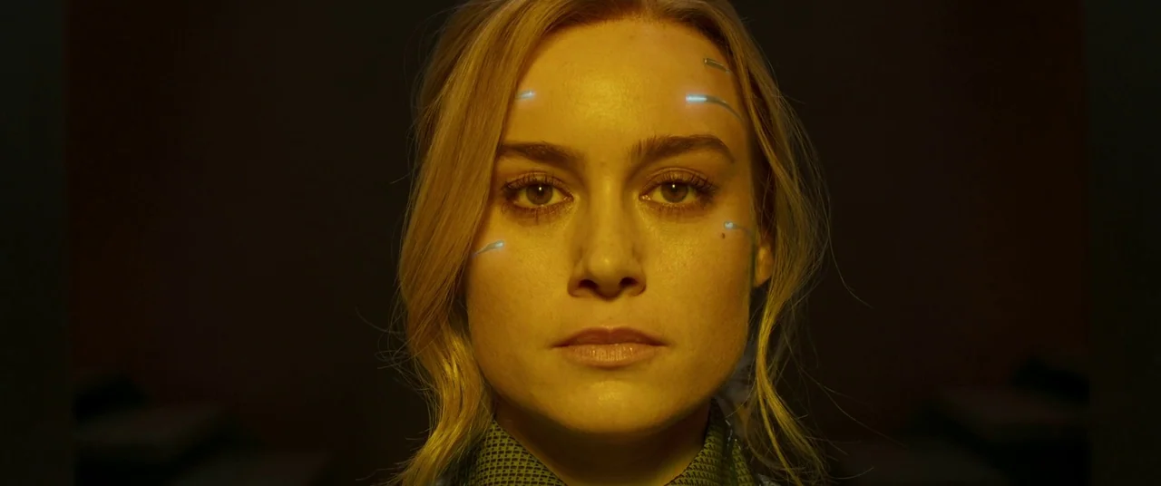 Captain Marvel (2019) movie still: closeup — A dimly lit room with muted dark tones and a stark amber color wash. A woman with glowing mechanical…