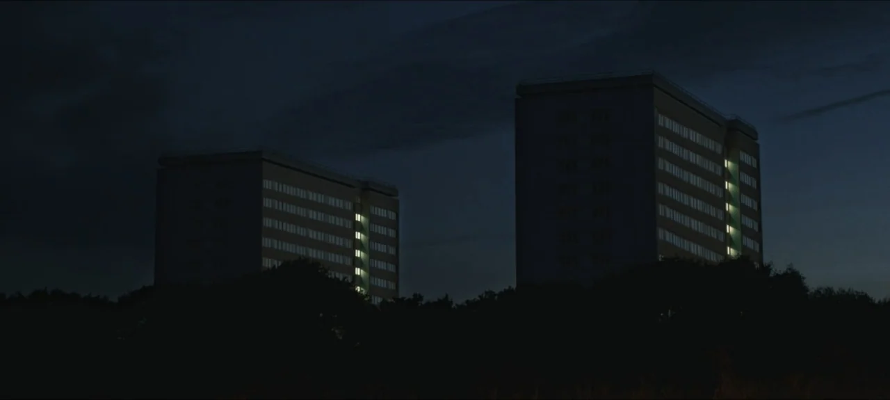 Containment (2015) movie still: establishing — Two identical multi-story apartment blocks rise above a dark silhouette of trees at night. Scattered…