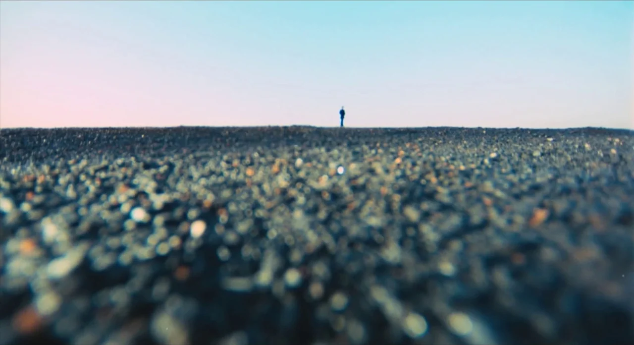 The Other Side of the Wind (2018) movie still: wide — A dark, pebbled beach stretches toward the horizon under a clear sky. A lone figure stands…