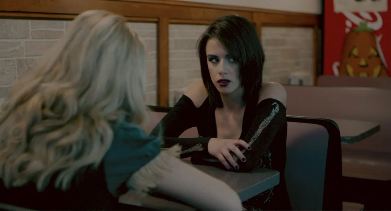 Terrifier (2018) movie still: medium — A diner booth features brown cushioned seating and a grey laminate table top. A dark-haired woman sits…