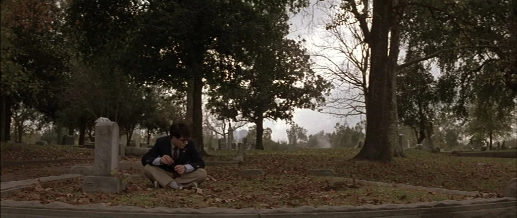 Rushmore (1998) movie still: wide — A quiet, overcast cemetery features numerous mature trees and worn stone headstones in rows. A young man wearing…