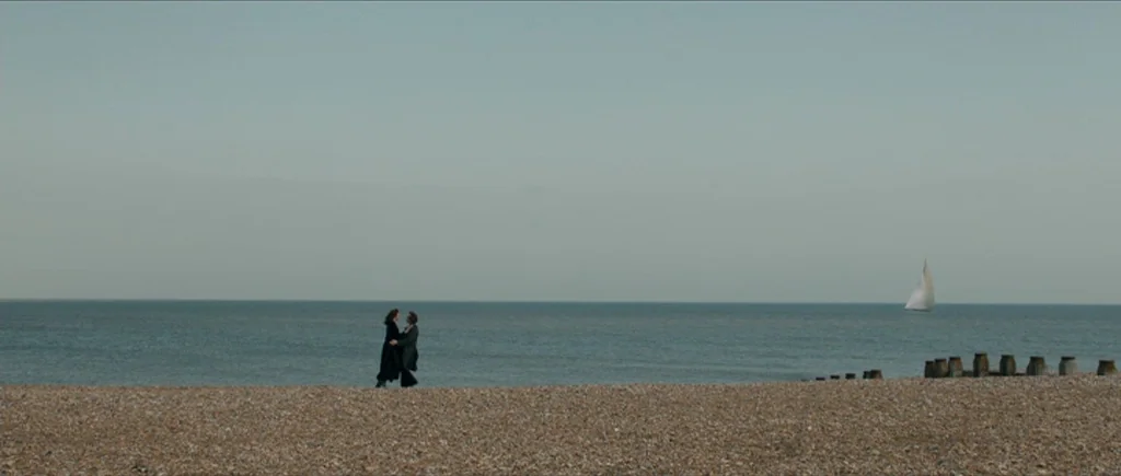 The Look of Love (2013) movie still: wide — A vast, grey pebble beach meets a calm, flat sea under a pale sky. Two figures in black coats stand on…