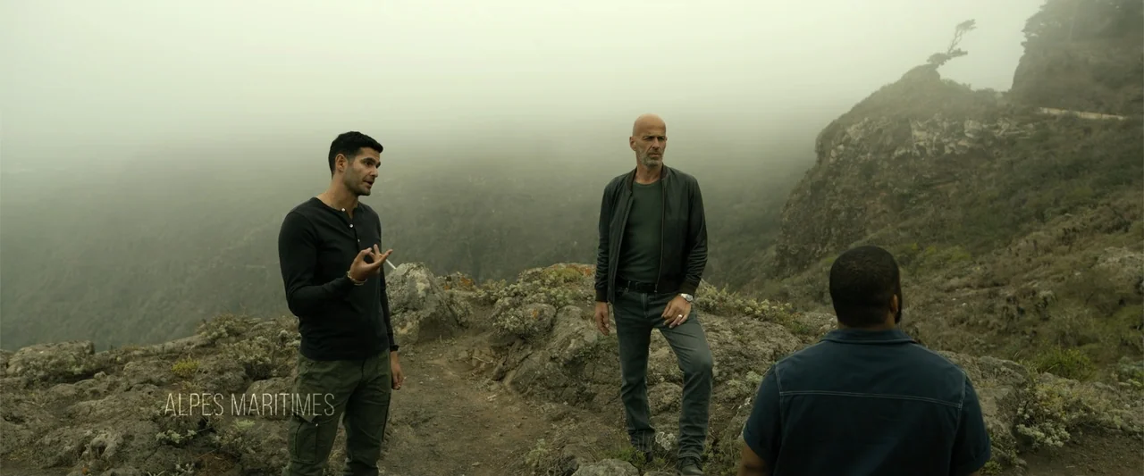 Den of Thieves 2: Pantera (2025) movie still: fullbody — A group of three men stands on a rocky, foggy mountain ledge overlooking a valley. The…