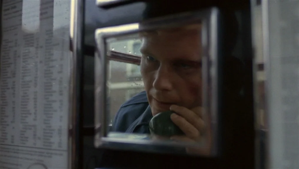 Performance (1970) movie still: closeup — A man stands inside a glass telephone booth with a printed directory visible. He holds the receiver…