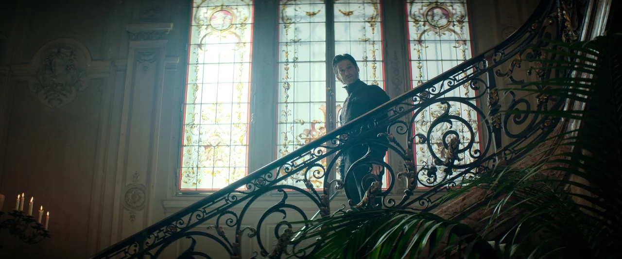The Count of Monte-Cristo (2024) movie still: fullbody — A man stands on a grand staircase before large, backlit stained-glass windows. Intricate…