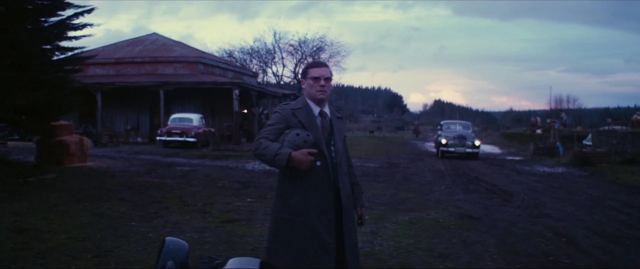 Neruda (2016) movie still: fullbody — A rural landscape at dusk features a small wooden structure with a gas pump. A man in a trench coat stands in…
