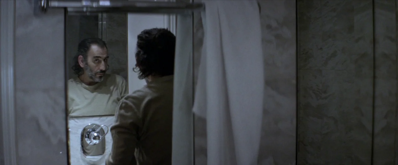 Chevalier (2015) movie still: medium — A dimly lit bathroom features marble wall tiling and a hanging white towel. A man stands facing a mirror,…
