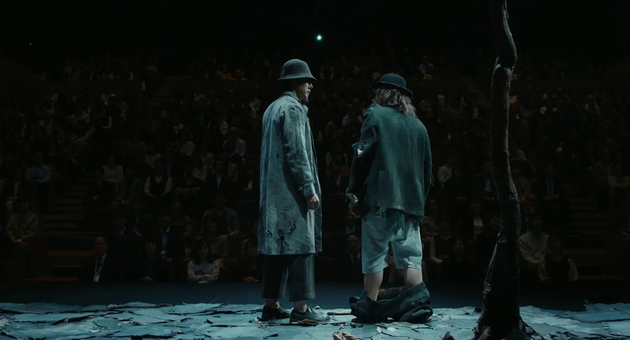 Drive My Car (2021) movie still: fullbody — A theatrical performance takes place on a sparse set featuring a single barren tree. Two men in tattered…