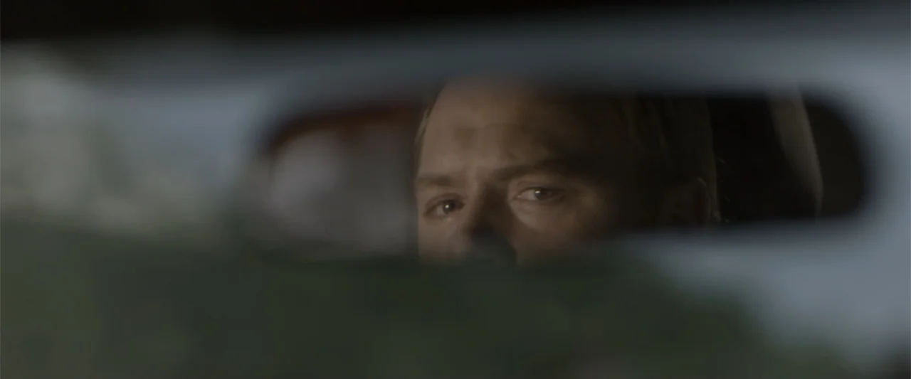 Radius (2017) movie still: closeup — A man's face is reflected in a car rearview mirror inside a vehicle cabin. The man is looking forward with a…