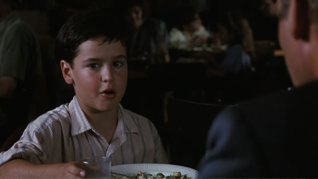 Presumed Innocent (1990) movie still: medium — A dark restaurant interior features a dimly lit dining area in the background. A young boy sits at a…