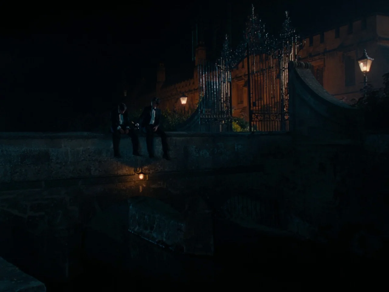 Saltburn (2023) movie still: wide — A stone bridge sits at night before a large ornate iron gate. Two men in tuxedos sit on the ledge of the bridge.