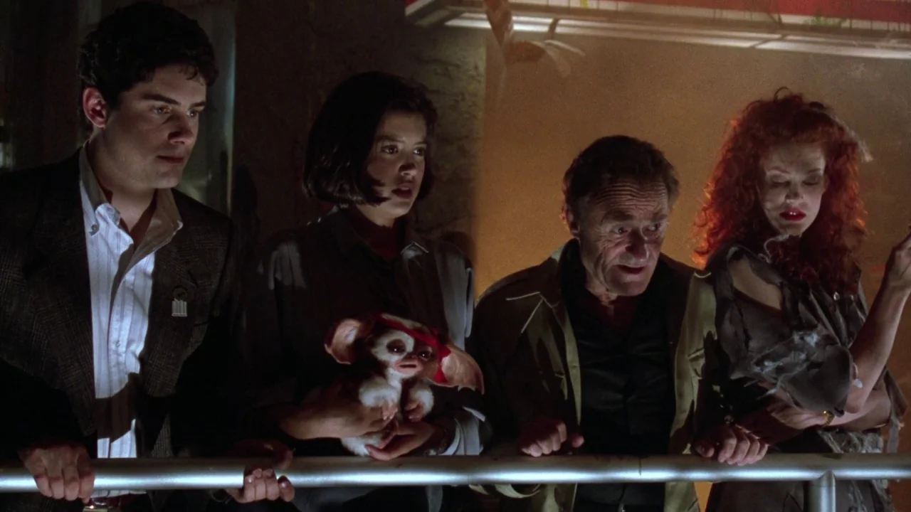 Gremlins 2: The New Batch movie still: medium — A dimly lit industrial room features a metal railing in the foreground. Four people stand behind the…