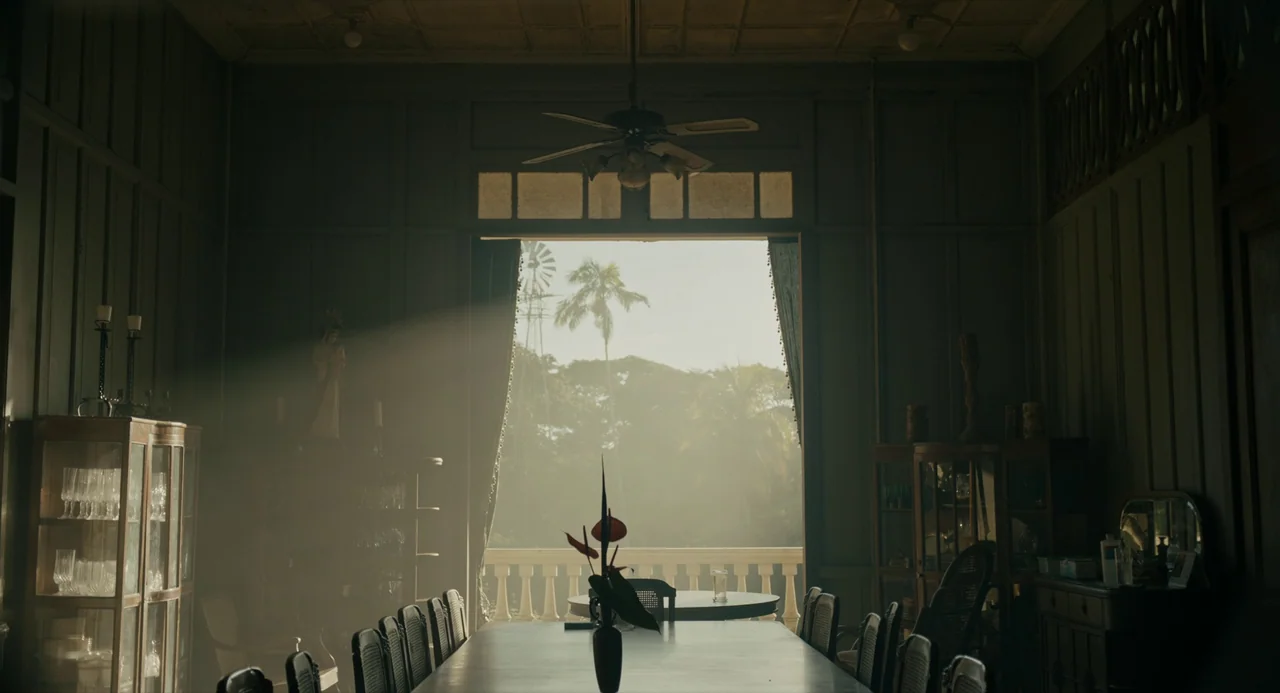 In My Mother’s Skin (2023) movie still: wide — A spacious dining room features dark wood-paneled walls and a long central table. Sunlight streams…