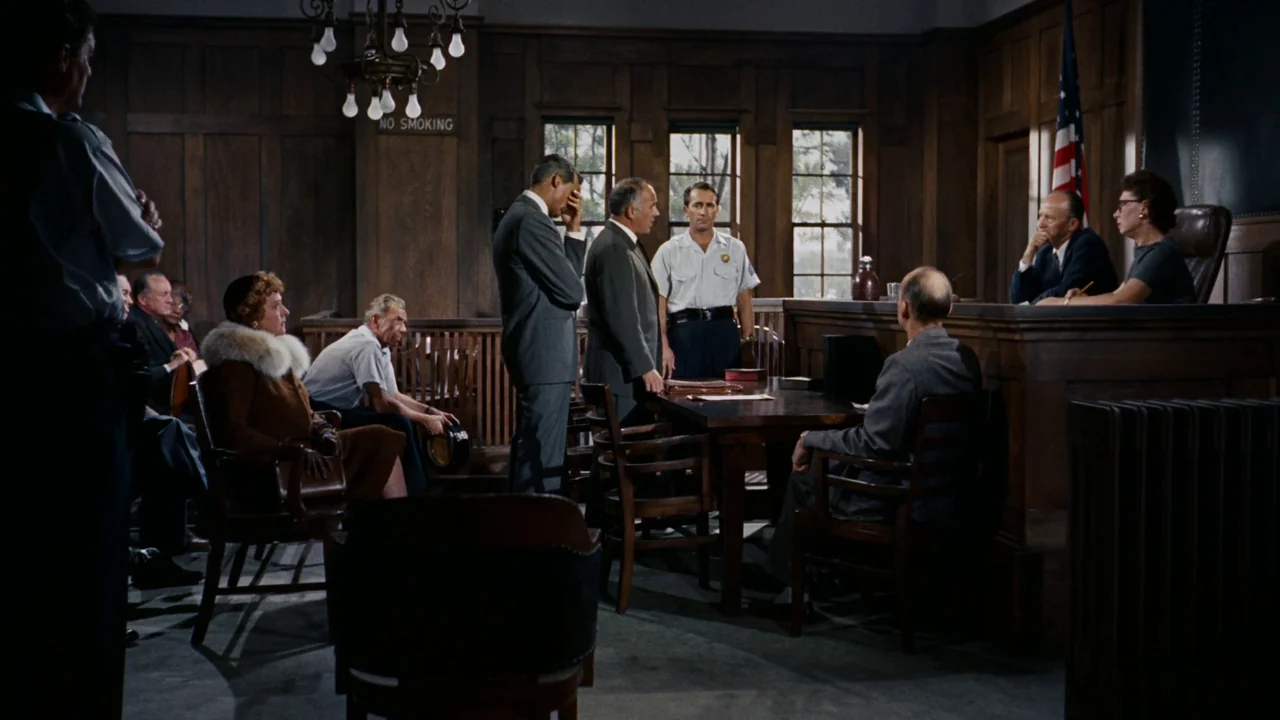 North By Northwest (1959) movie still: wide — A courtroom features dark wood paneling, windows with outdoor light, and a US flag. Several people are…