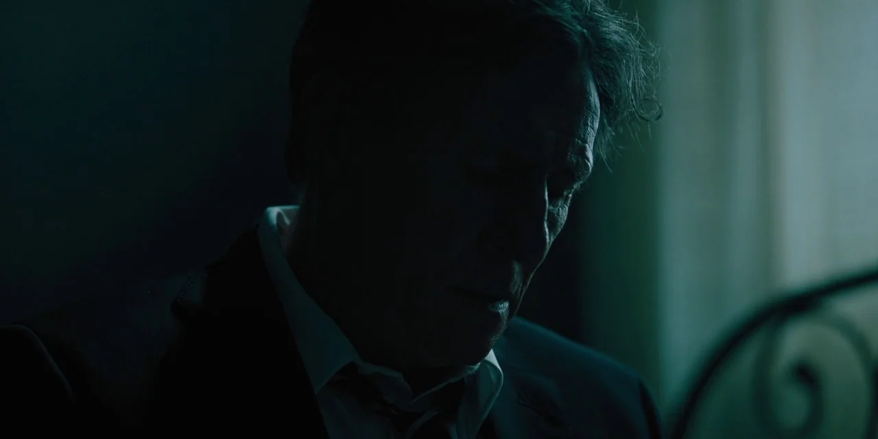 Hereditary (2018) movie still: closeup — A dimly lit interior space with a dark, indistinct background. A mature man in a suit looks down with his…