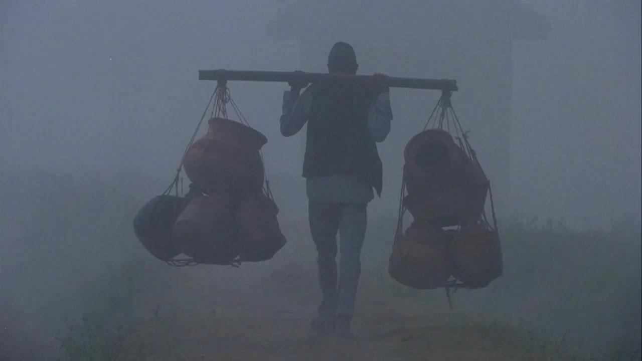 Powaqqatsi (1988) movie still: fullbody — A man walks away along a narrow dirt path shrouded in thick grey fog. He carries a long bamboo pole across…