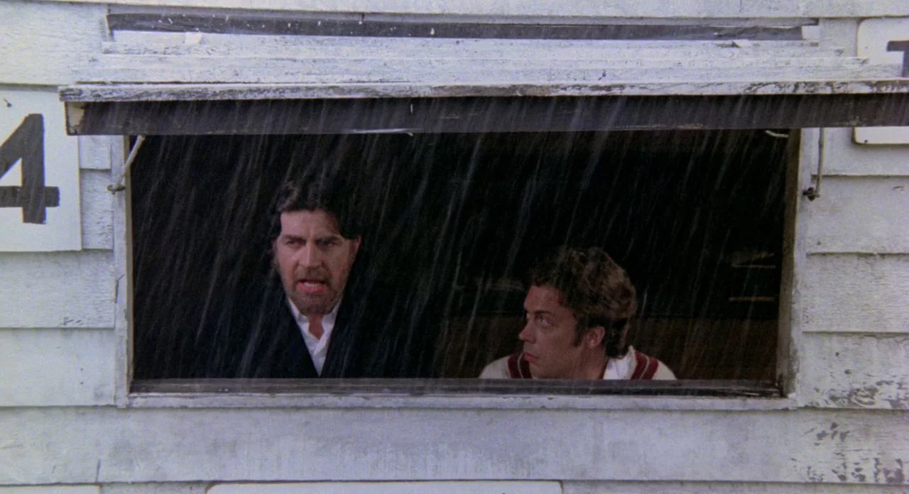 The Shout (1978) movie still: medium — Two men stand inside a weathered white wooden structure during a heavy rainstorm. They look outward through a…
