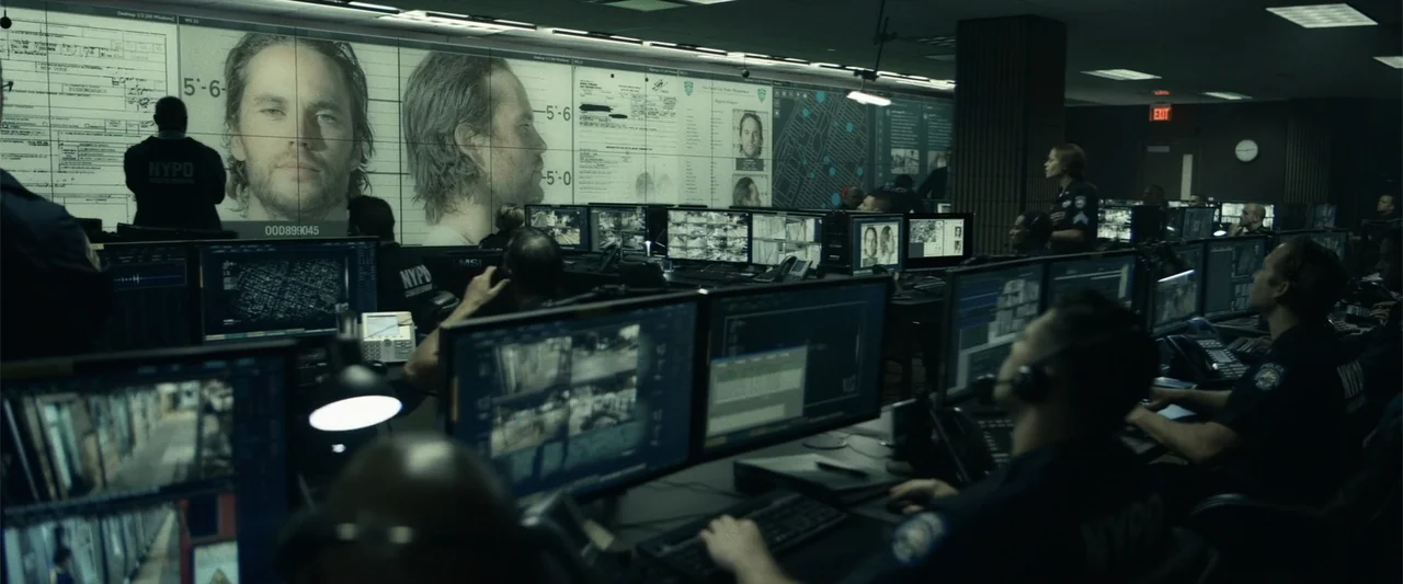 21 Bridges (2019) movie still: wide — A high-tech surveillance room features rows of glowing computer terminals and a massive wall display. Multiple…
