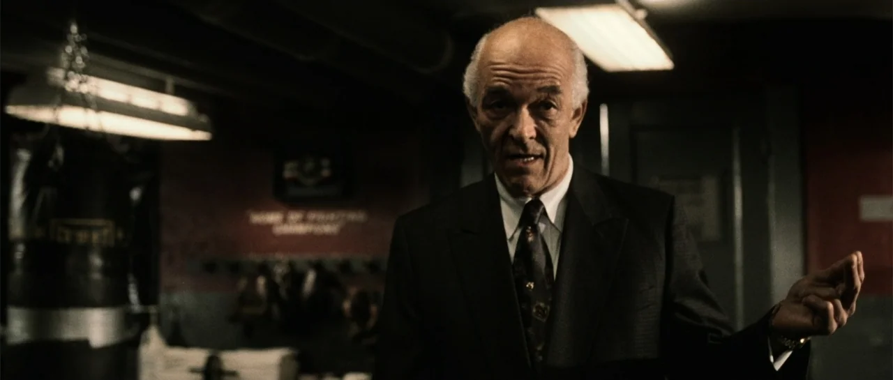 Daredevil (2015) movie still: medium — A dimly lit indoor space contains a dark punching bag in the foreground. An older man in a suit stands facing…