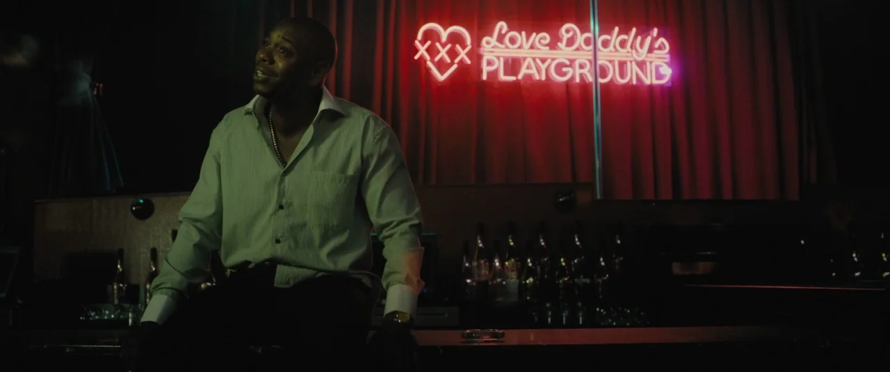Chi-raq (2015) movie still: medium — A dimly lit club interior features a prominent red neon sign on the back wall. A man sits on the edge of the…