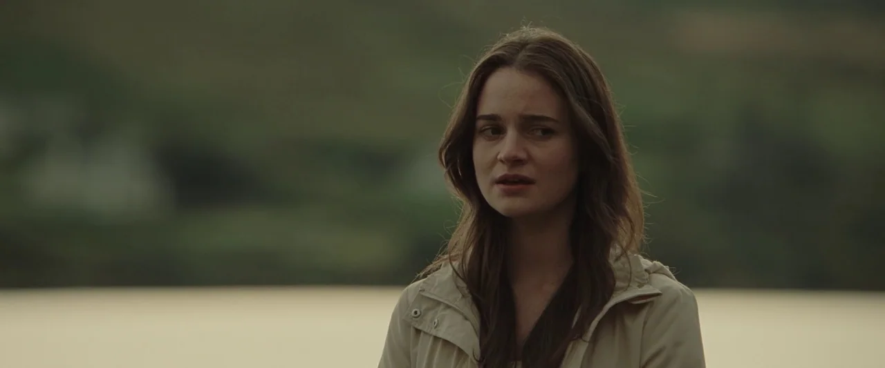 God’s Creatures (2022) movie still: medium — A woman stands in front of a blurry, verdant landscape and a calm body of water. The subject is gazing…
