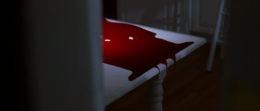 American Beauty (1999) movie still: closeup — A white dining table is covered in a large, thick pool of deep red blood. The liquid spills over the…