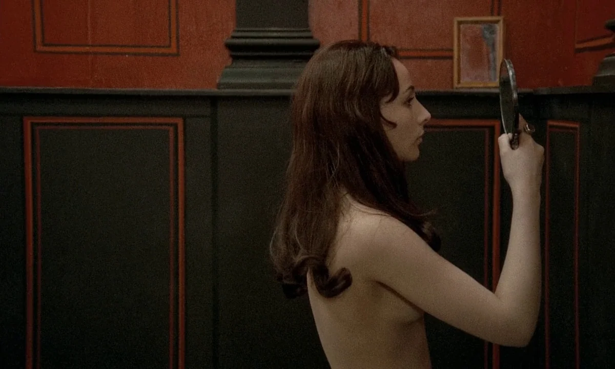 Immoral Tales (1974) movie still: medium — An interior space with dark charcoal wainscoting and deep red panels featuring thin borders. A woman…