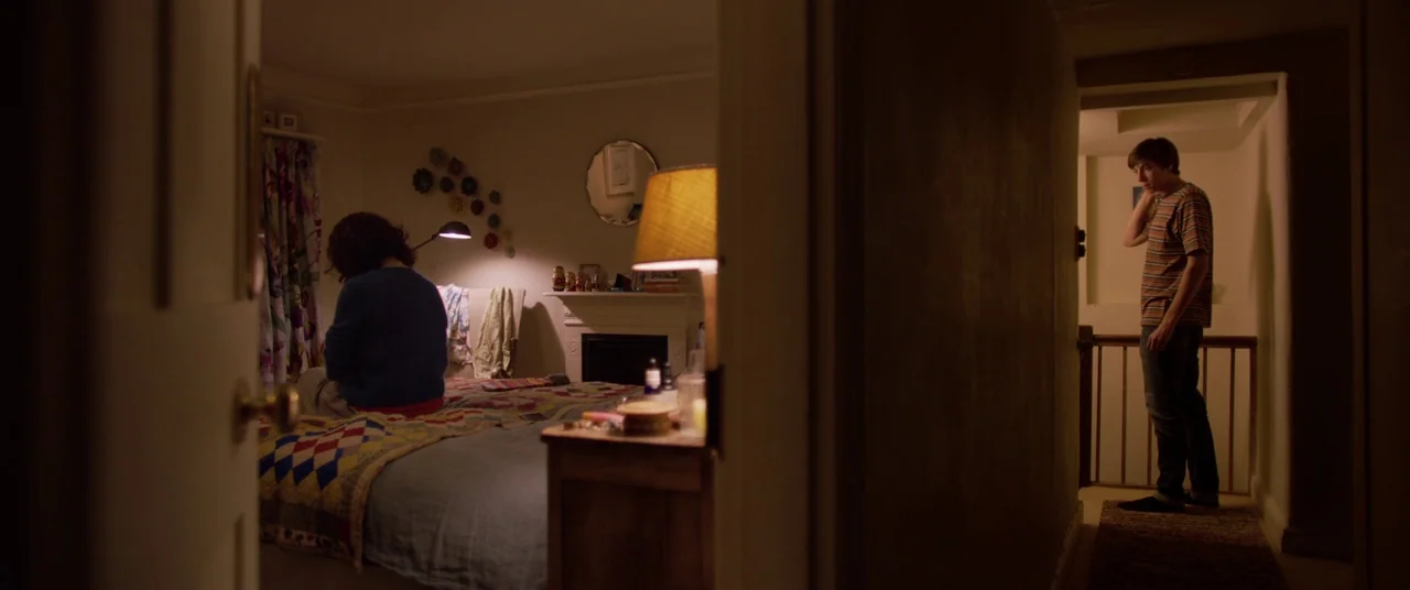 Yesterday (2019) movie still: wide — A dimly lit room with a patterned quilt on a bed is visible through a doorway. A young man stands in a shadowed…