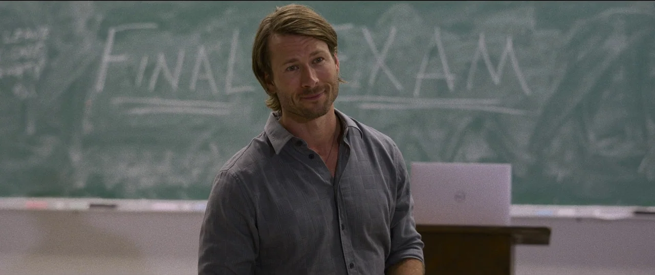 Hit Man (2023) movie still: medium — A man stands before a chalkboard with the words Final Exam written in chalk. He looks toward the camera with a…