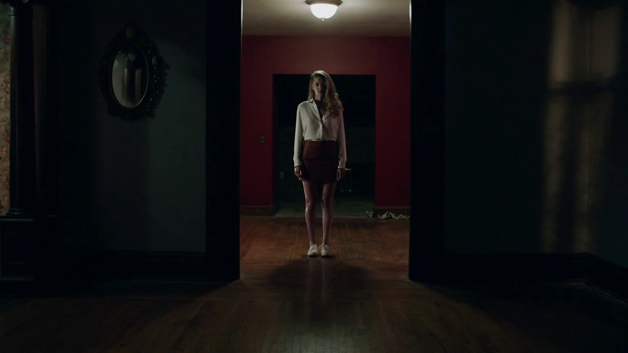Girl on the Third Floor (2019) movie still: fullbody — A dark, dimly lit domestic hallway with hardwood floors and a deep red back wall. A young…