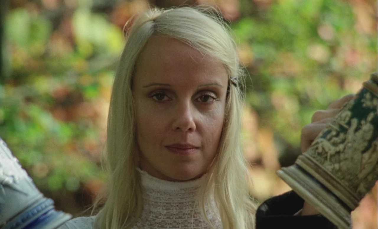Mahler (1974) movie still: closeup — A woman stands outdoors against a background of green foliage. She holds a patterned ceramic beer stein in her…