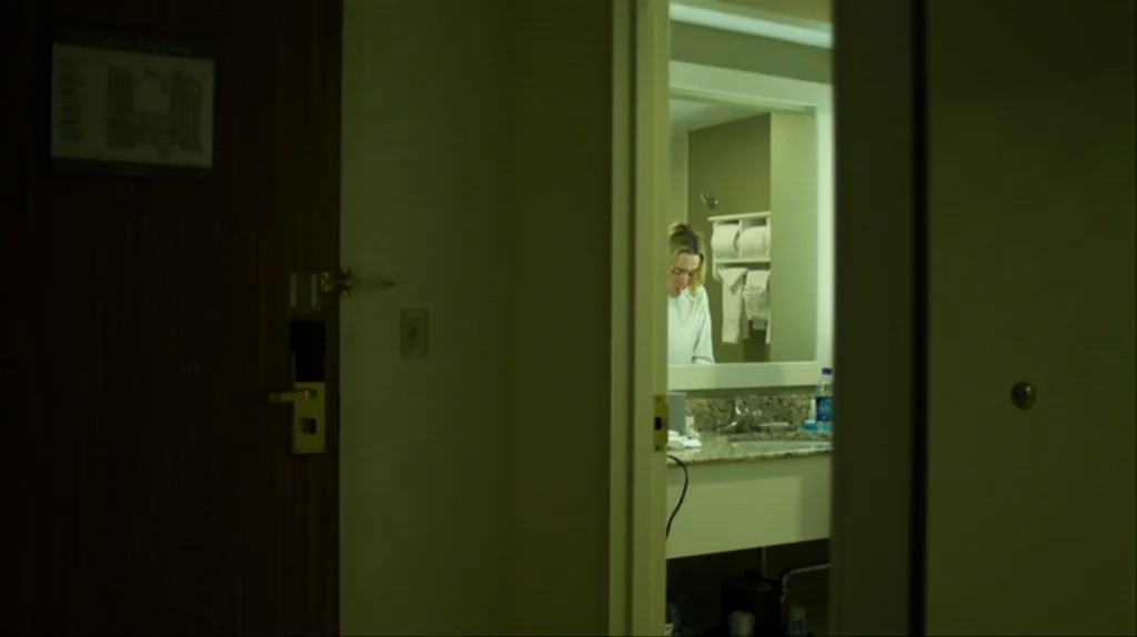 Contagion (2011) movie still: wide — A dark hotel room is seen through a door frame looking into a brightly lit bathroom. A woman stands at the…
