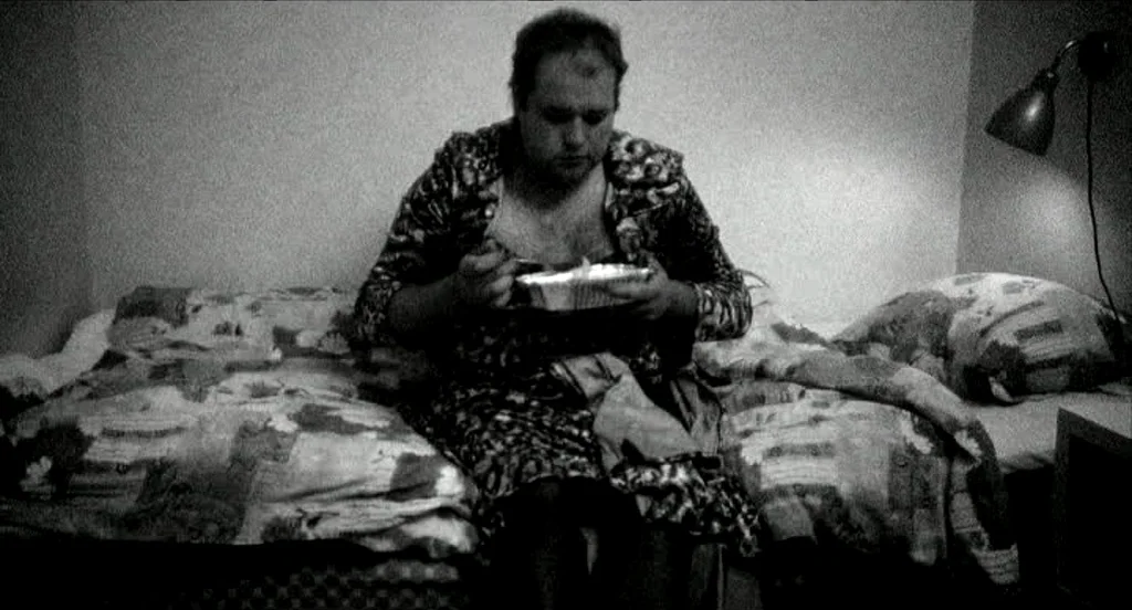 Container (2006) movie still: medium — A sparsely furnished room contains a single bed with a patterned comforter and pillows. The man sits on the…