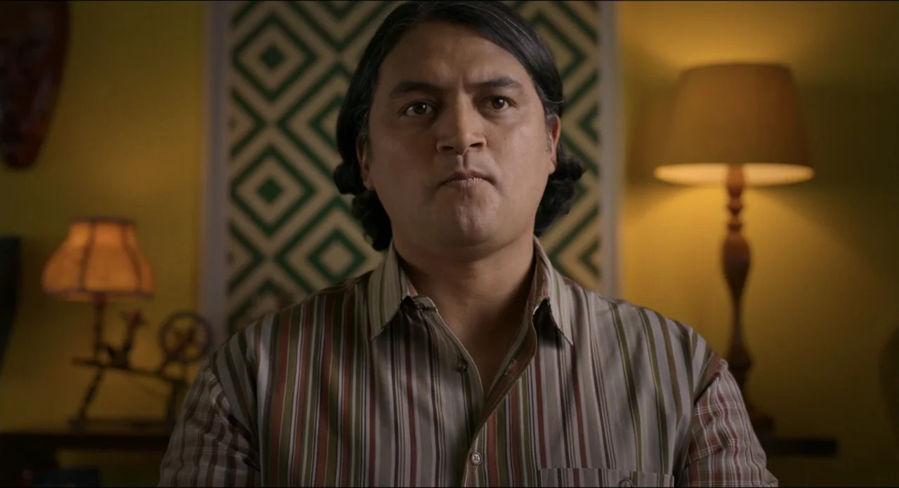 The Beauty and the Moody (2022) movie still: medium — A man sits in front of a patterned wall with two table lamps flanking him. The subject looks…