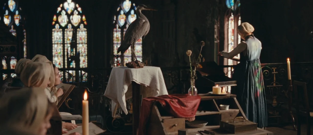 Madame Bovary movie still: wide — A dark space with multiple stained glass windows and several wooden desks with candles. Students in period…