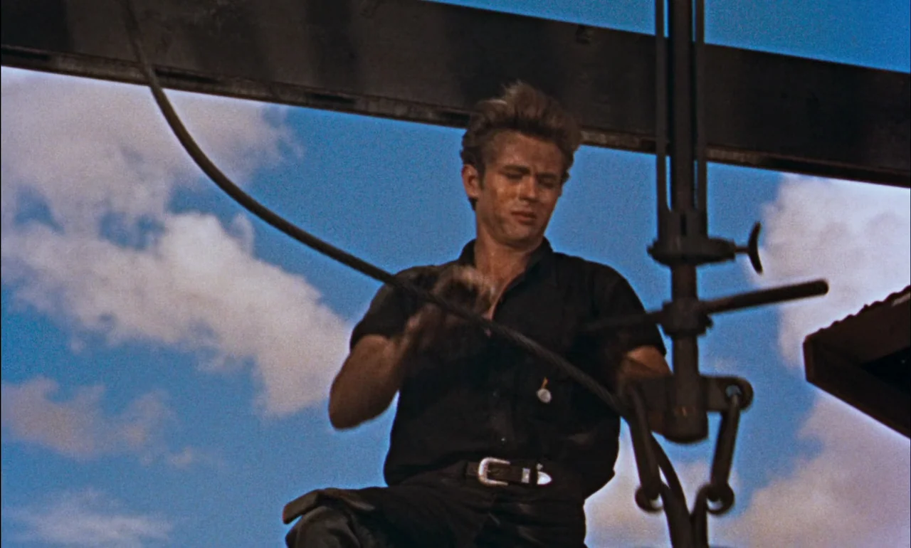 Giant (1956) movie still: medium — A man works on industrial equipment set against a bright, cloud-filled sky. He handles a thick metal cable while…