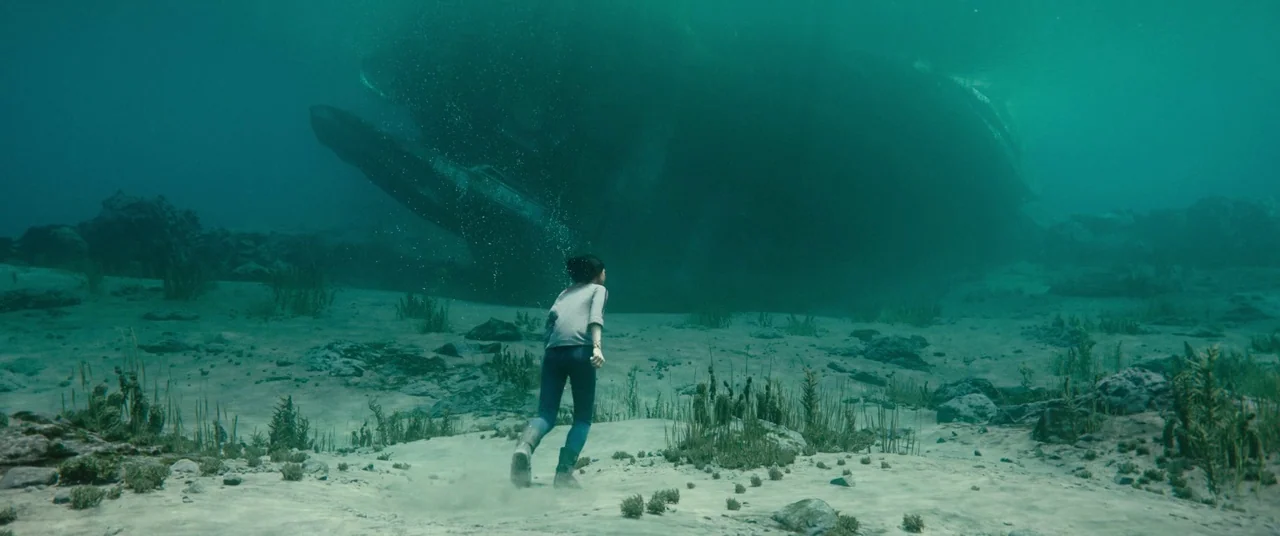 Alita: Battle Angel movie still: wide — A sandy seabed with sparse patches of aquatic vegetation and small rocks. A young woman runs across the…