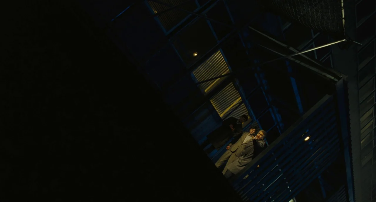 The Double (2013) movie still: wide — Three men stand on an elevated metal platform at night near a modern building. They look outward while harsh…