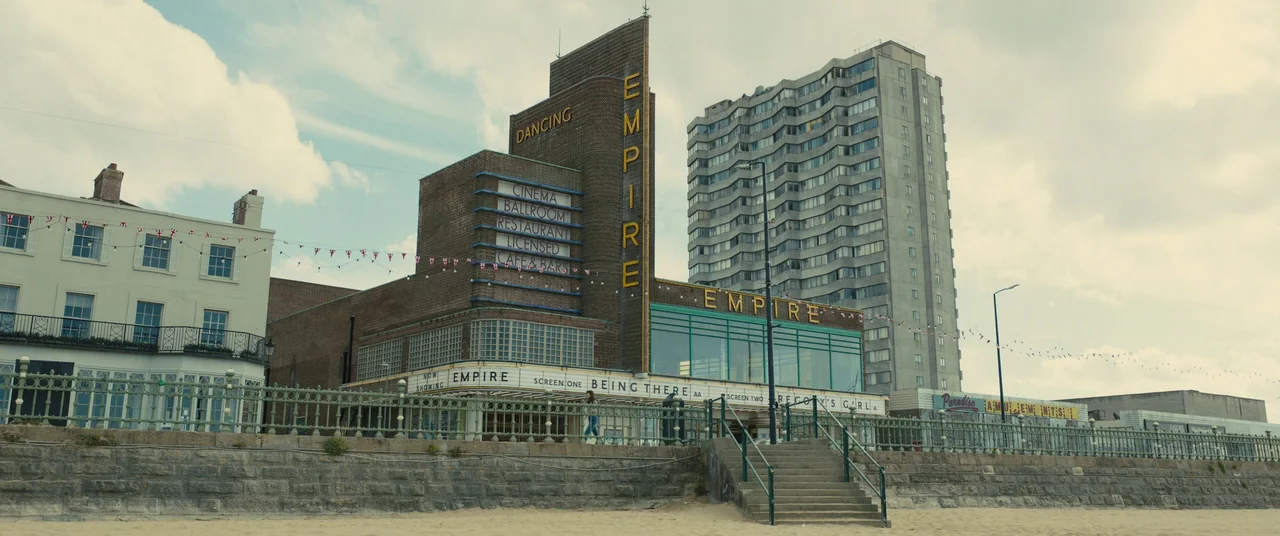 Empire of Light (2022) movie still: wide — A seaside promenade features an art deco theater building with prominent yellow signage. Adjacent to the…