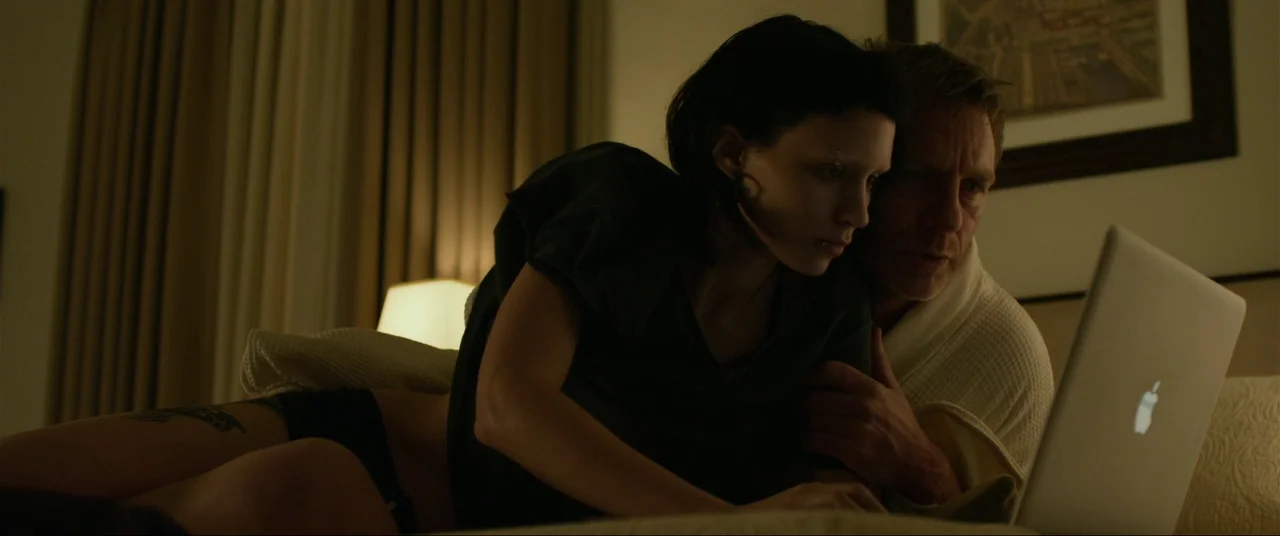 The Girl With The Dragon Tattoo (2011) movie still: medium — A dimly lit hotel room with a beige wall featuring a framed print behind the bed. A man…