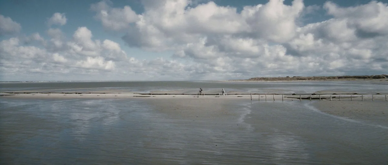 Womb (2010) movie still: wide — Expansive wet tidal flats stretch toward a cloudy horizon under a vast sky. Two individuals traverse a thin wooden…