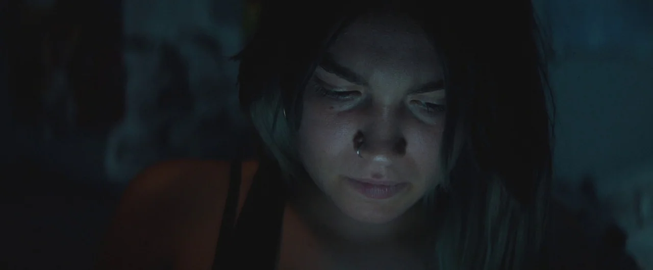Poser (2021) movie still: closeup — A person is situated in a dark interior space with no other visible objects. The individual looks downward…
