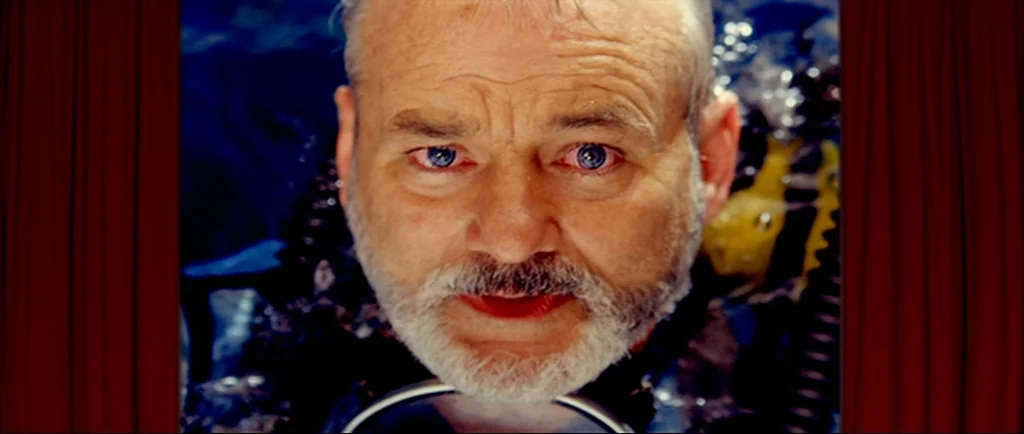 The Life Aquatic With Steve Zissou (2004) movie still: closeup — An older man submerged in blue water is framed by vertical red stage curtains. The…
