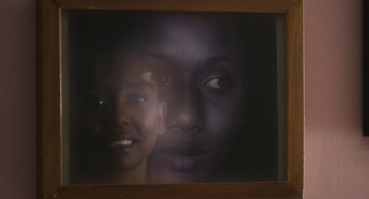 Zombi Child (2019) movie still: closeup — A square wooden picture frame hangs against a plain, dimly lit wall surface. The glass surface displays a…