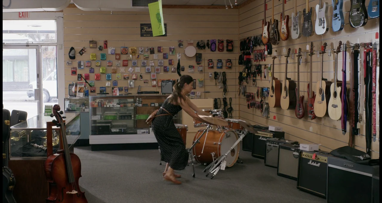 One True Loves (2023) movie still: fullbody — A music retail space with guitar racks on a slatwall and a display case. A woman in a patterned dress…